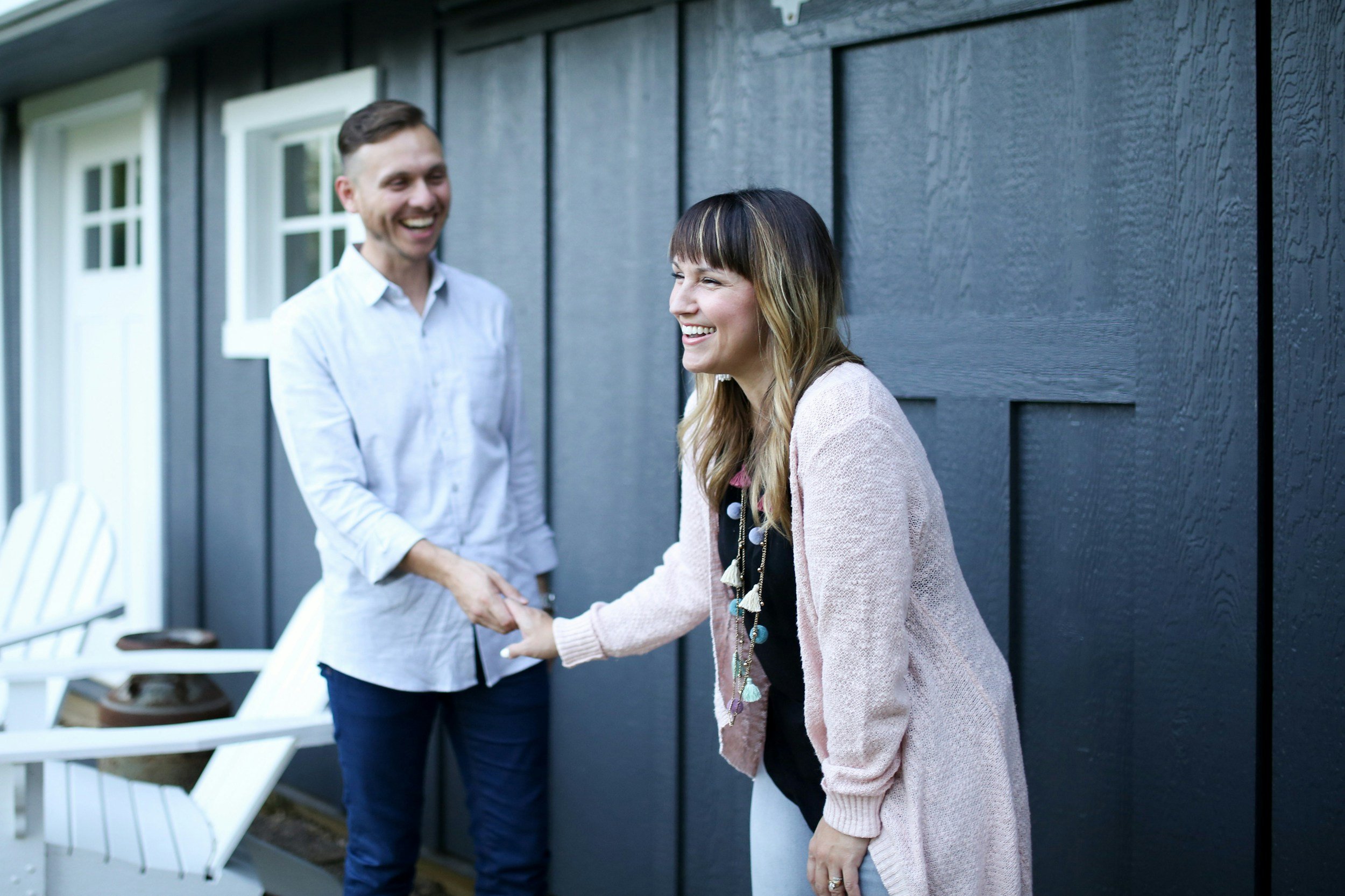 A man and a woman are smiling and shaking hands outside a house with dark wood siding. The woman is wearing a pink cardigan and a colorful necklace, and the man is wearing a white shirt.