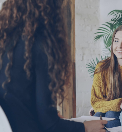 Two women having a conversation indoors, one with curly hair and the other with long straight hair, sitting in front of a plant and a white brick wall.
