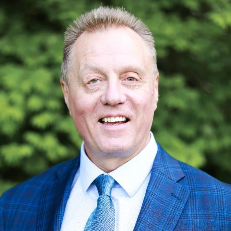A middle-aged man smiling outdoors in front of green trees, dressed in a blue plaid suit, light blue shirt, and matching blue tie.