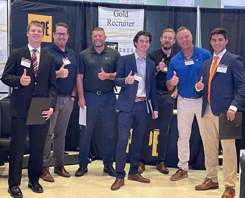 Group of seven men at a career fair or conference, standing in front of a banner titled 'Gold Recruiter,' all giving thumbs-up signs while dressed in business or business casual attire.