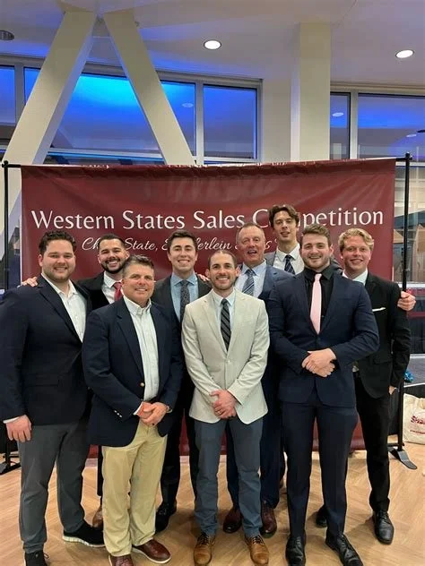 Group of ten men in formal attire standing in front of a red banner that reads "Western States Sales Competition" at an indoor event.