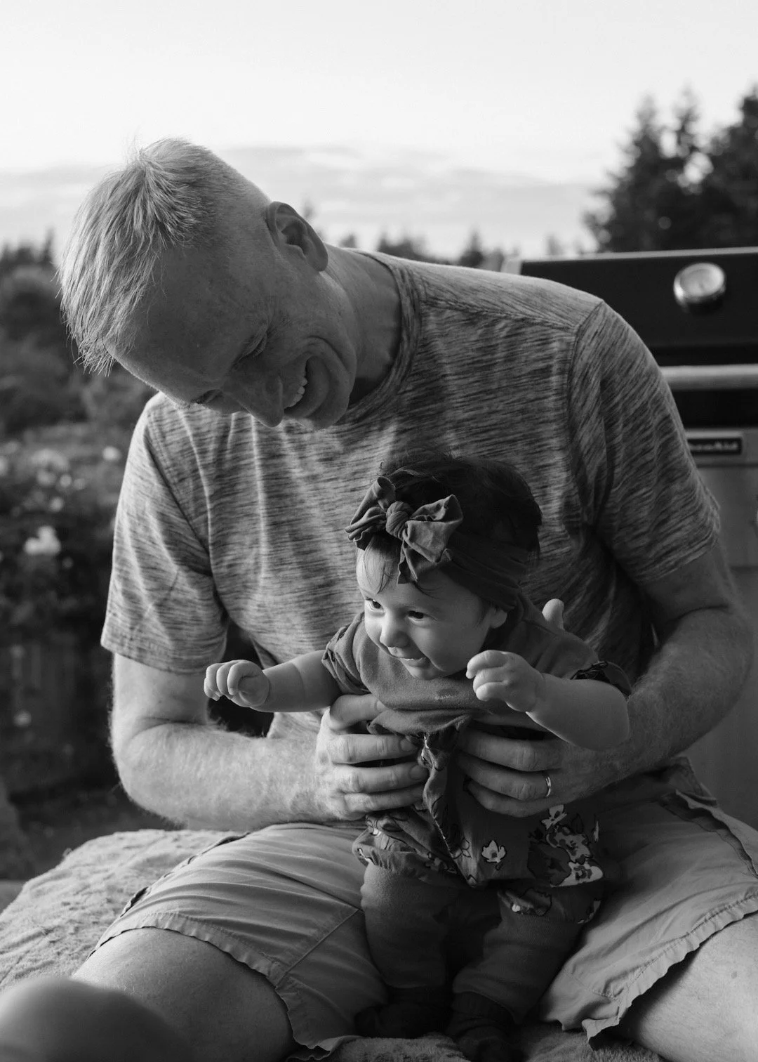 An older man with light hair and a short-sleeved shirt holding a young girl with a bow in her hair, both smiling and enjoying a moment outdoors.