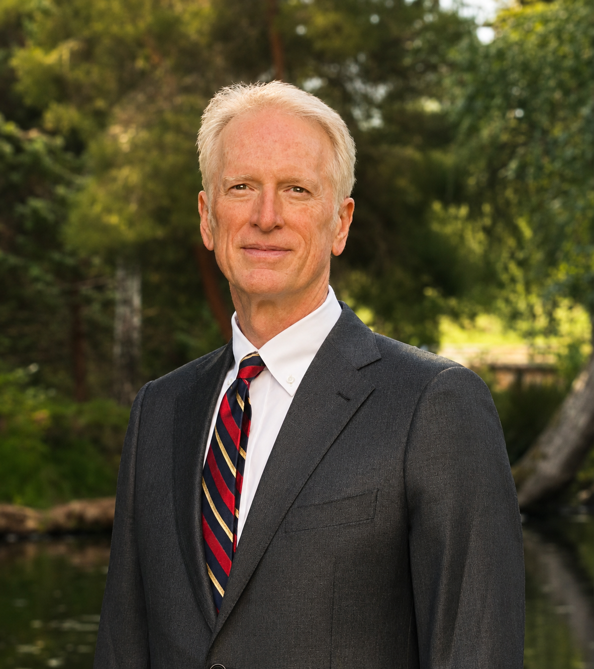A middle-aged man with light skin, blond hair, and a slight smile, dressed in a dark suit, white shirt, and a striped tie, standing outdoors with green trees and a body of water in the background.