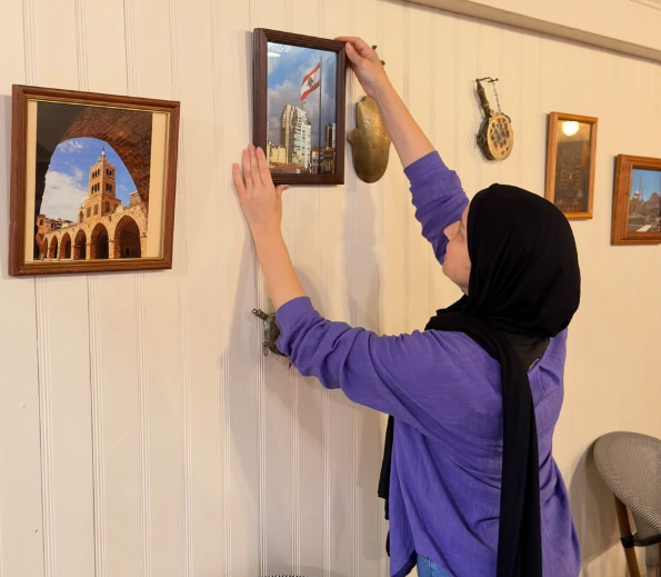 A woman wearing a black headscarf and purple sweater hanging a small framed picture on a wall decorated with other framed artworks and musical instruments.