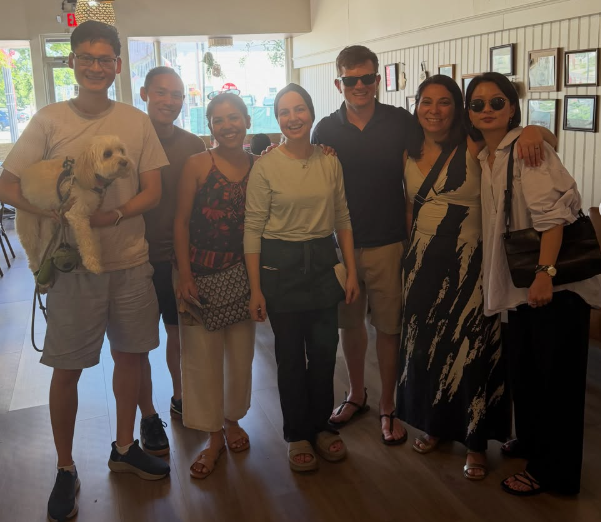 Group of seven people standing inside a bright room, smiling for the photo, with one person holding a small white dog.