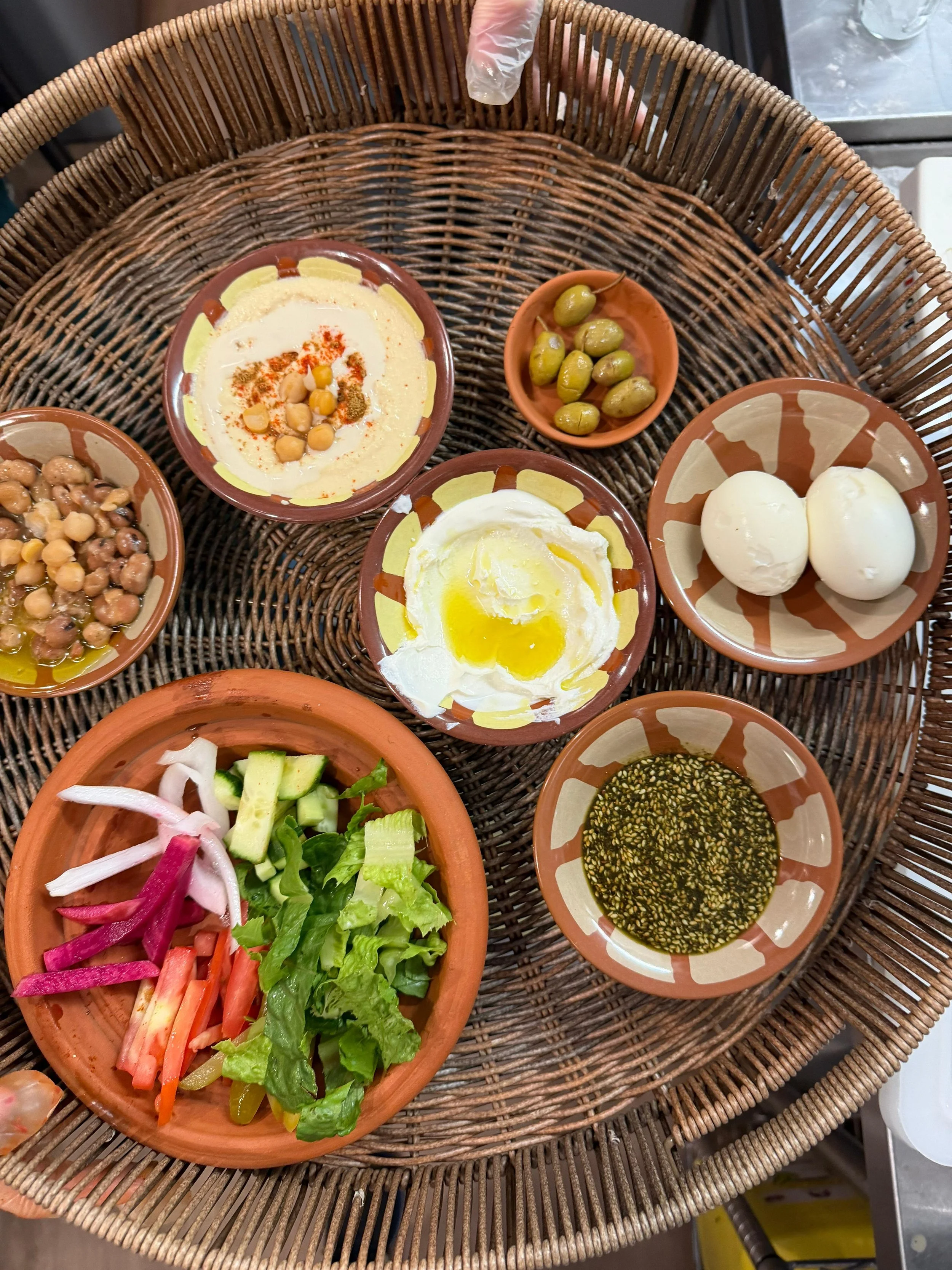 A variety of small bowls with condiments, salads, and sauces on a round wicker table. Includes a bowl of chickpeas, a bowl with eggs, a bowl of salad vegetables, and bowls of creamy dips, herbs, and sauces.