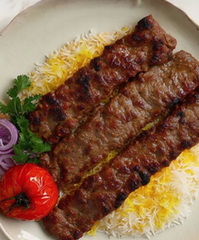 Two grilled meat kebabs on a plate with rice, cherry tomato, sliced red onion, and cilantro garnishes.