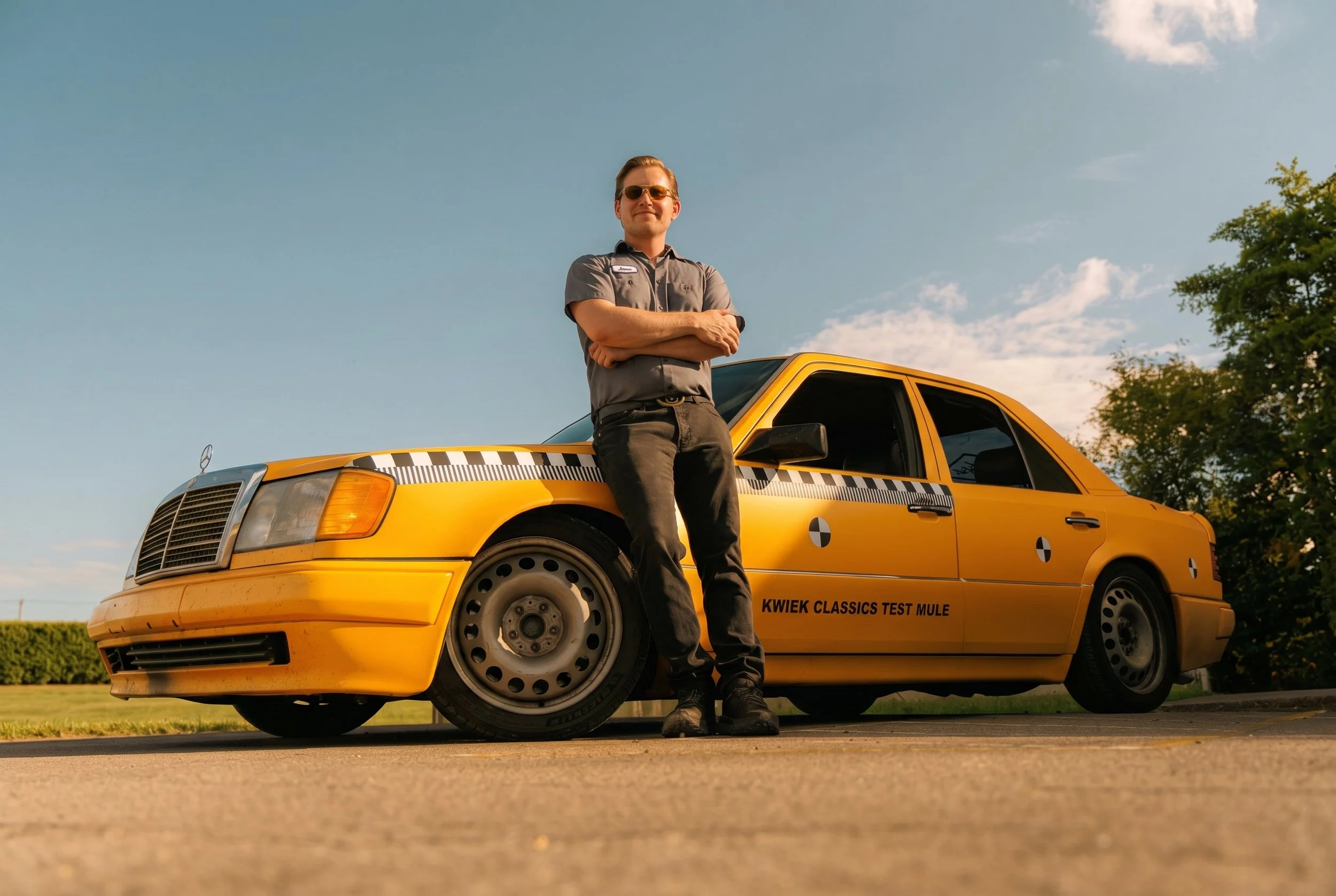 Matt Kwiek wearing sunglasses and a gray mechanic uniform stands with arms crossed in front of a yellow test mule car marked "Kwik Classics Test Mule," outdoors on a road with green trees and blue sky in the background.