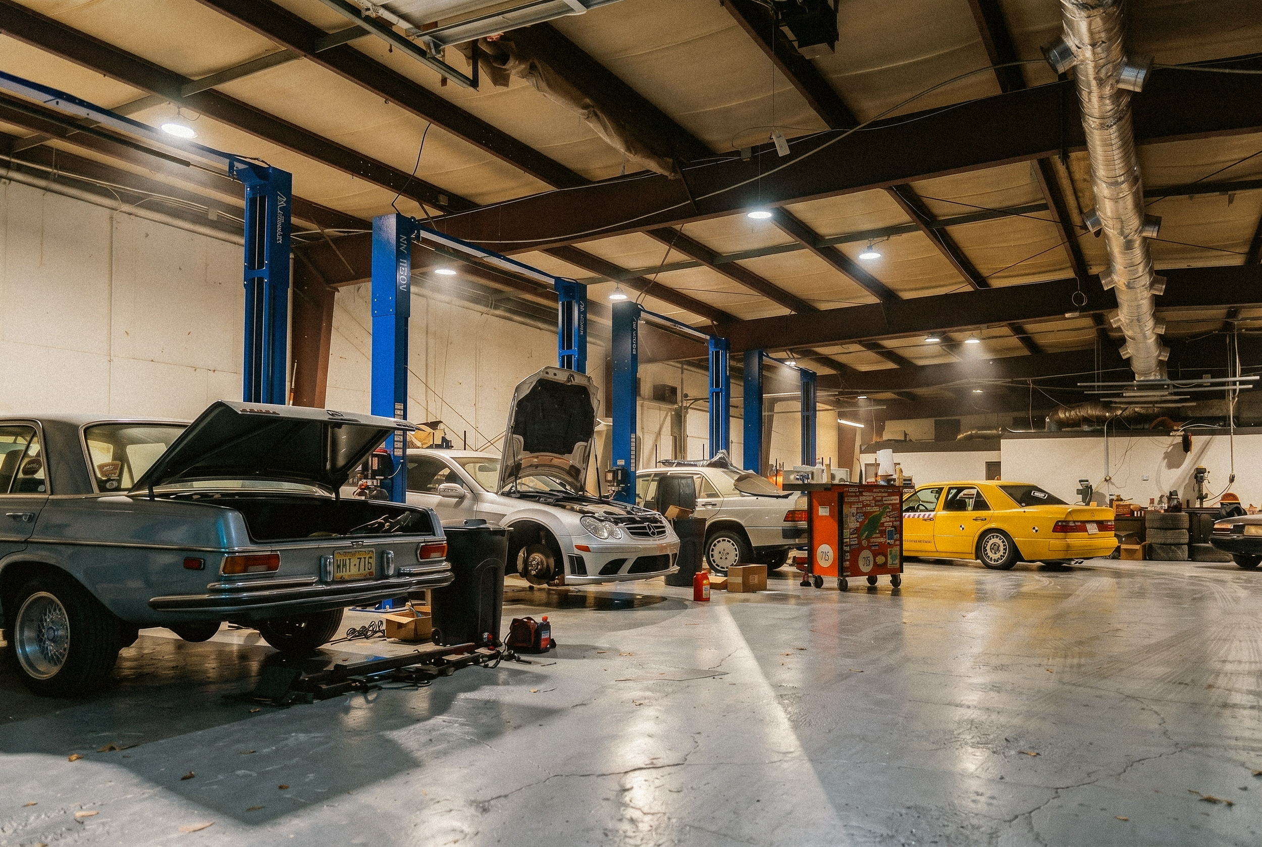 Inside KWIEK Classics' car repair shop with multiple vehicles being serviced. There are various tools, equipment, and car parts scattered around. The ceiling has exposed beams, ducts, and industrial lighting.