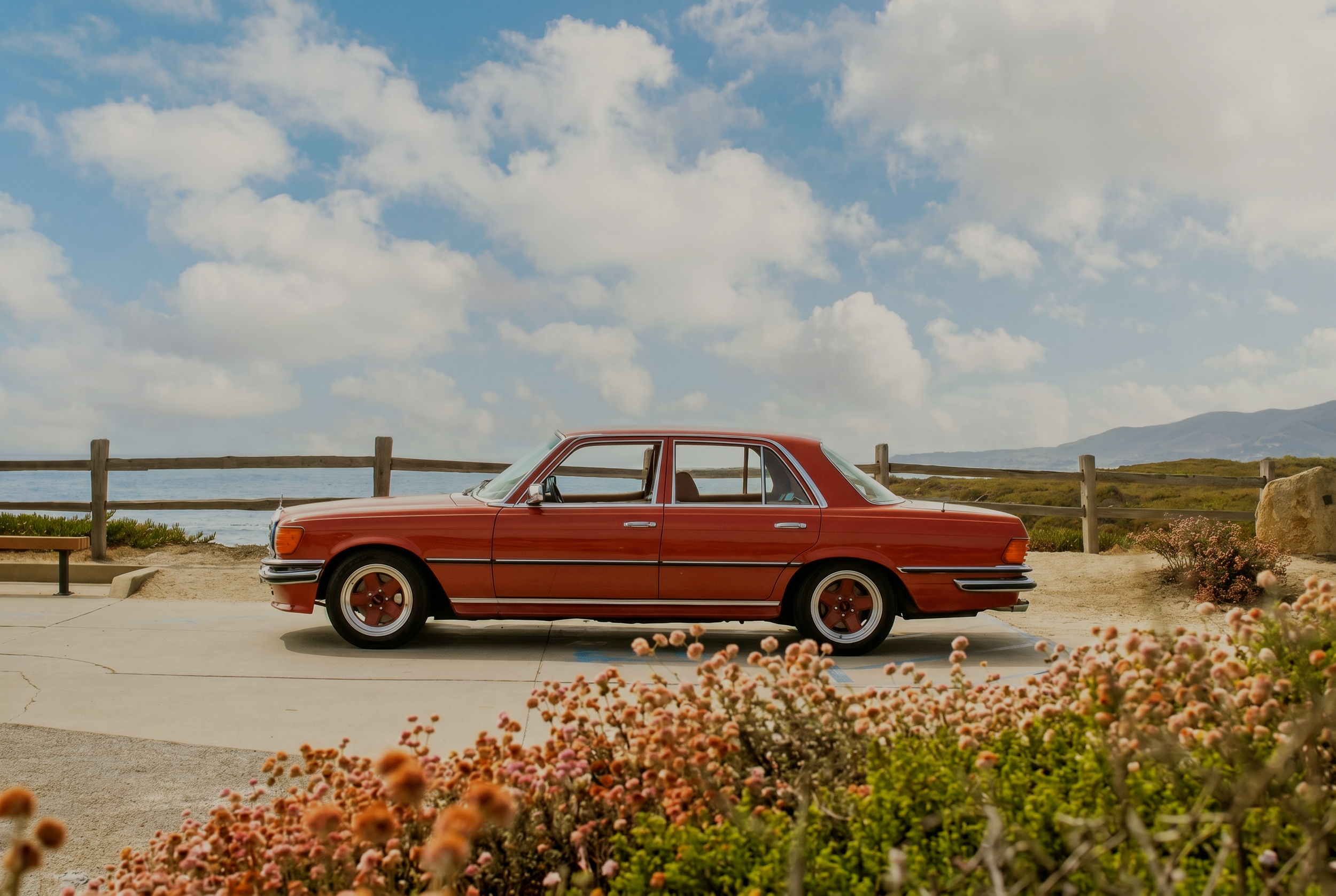 A red vintage car parked by a seaside park with a wooden fence, colorful flowers, and a cloudy sky.