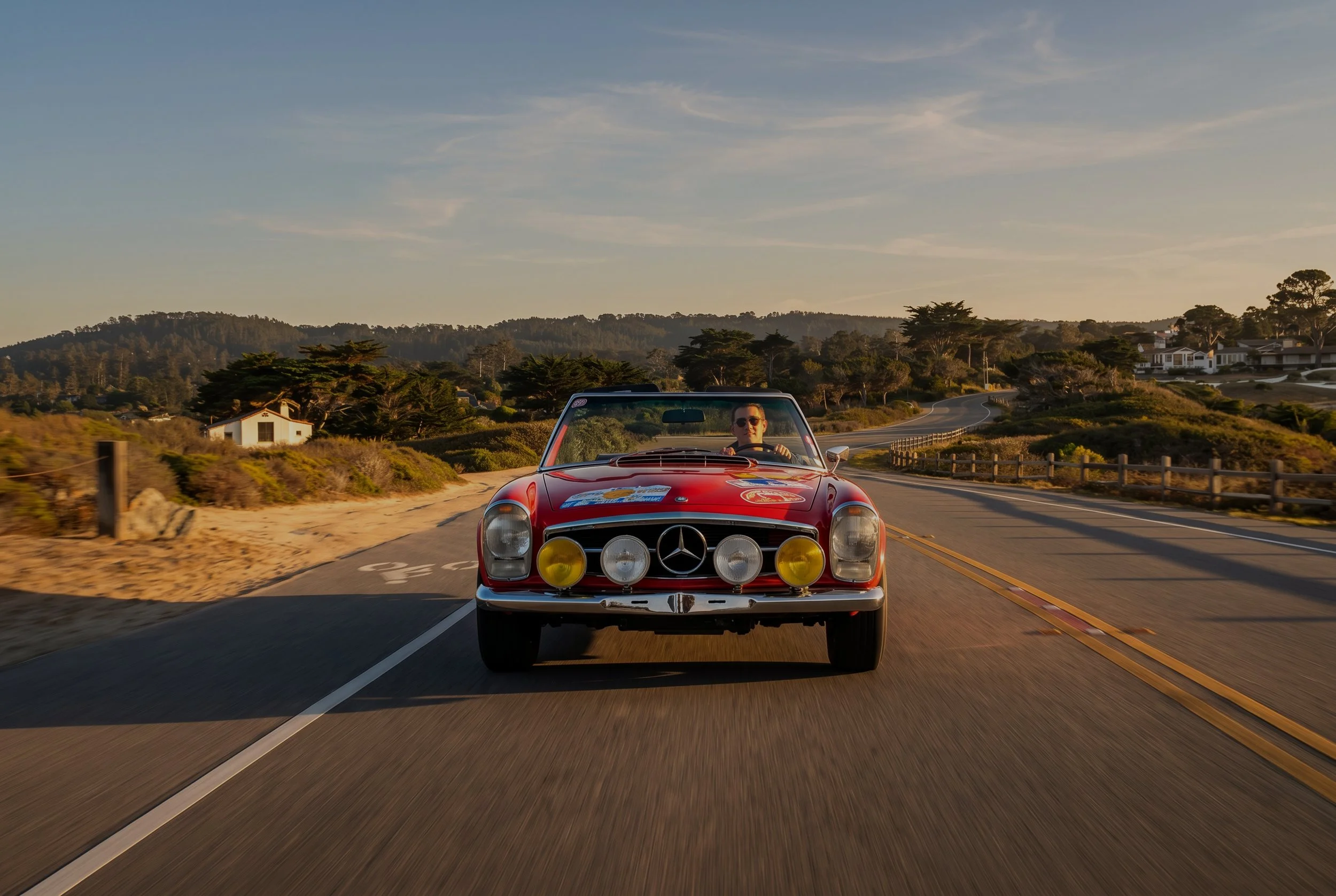 A man wearing sunglasses driving a red vintage Mercedes-Benz convertible with yellow fog lights on a scenic coastal road during sunset.