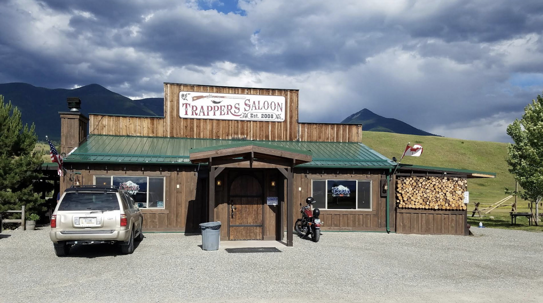 A rustic wooden building named Trappers Saloon with a green metal roof, American and state flags, parked vehicles including an SUV and a motorcycle, logs stacked on the side, and a backdrop of green hills and mountains under a cloudy sky.