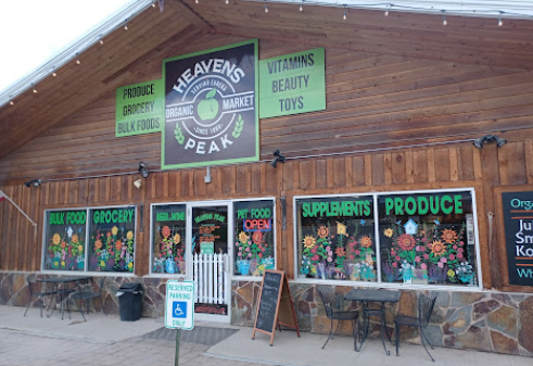 Exterior of Heaven's Peak Market with signs for produce, bulk foods, organic foods, groceries, vitamins, beauty products, and toys, featuring colorful window displays of flowers.