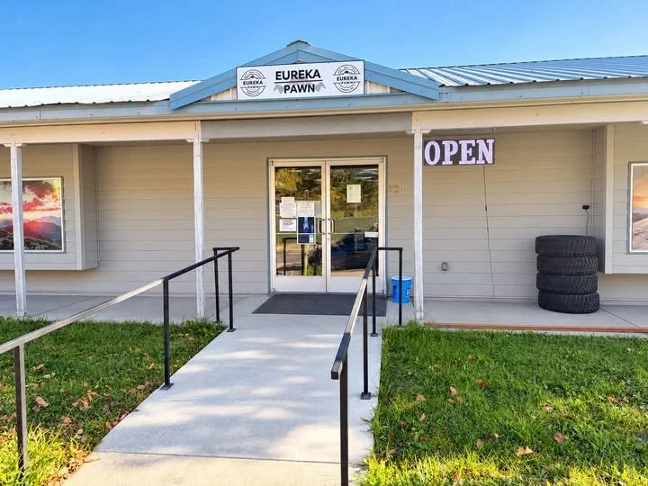 Store with a sign saying 'the Feed Bin' and an 'OPEN' sign, bags of animal feed stacked outside, large black tubing, and a gray metal building with a garage door.