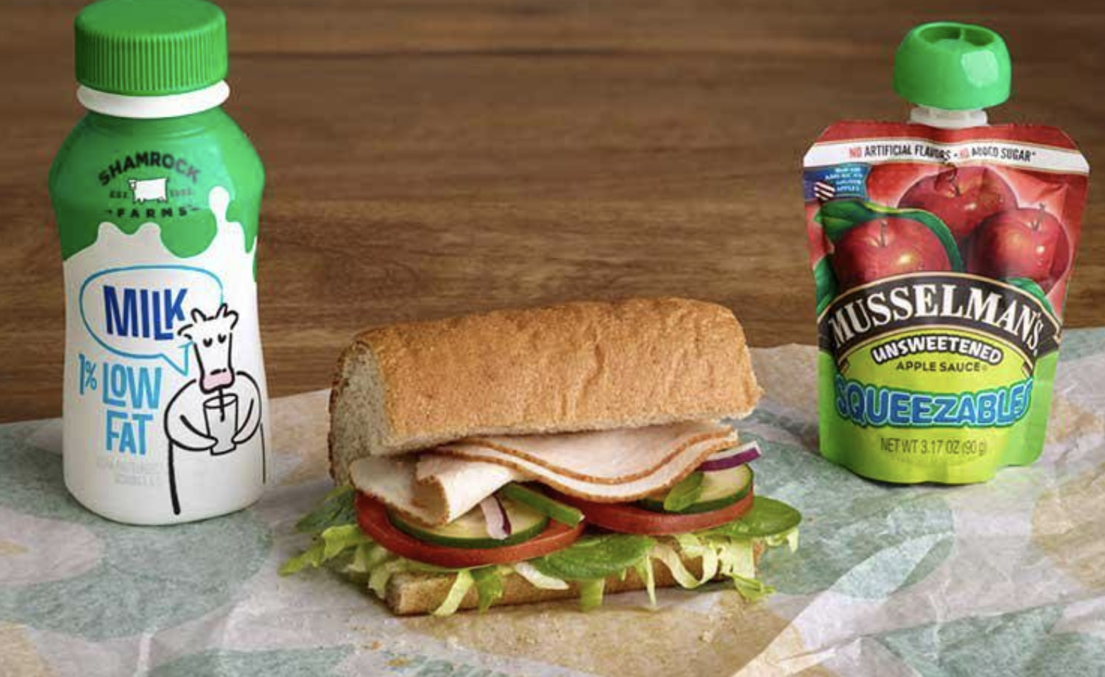 A turkey sandwich with lettuce, tomato, cucumber, and turkey slices on a sandwich bun, with a carton of milk and a pouch of apple sauce on either side.