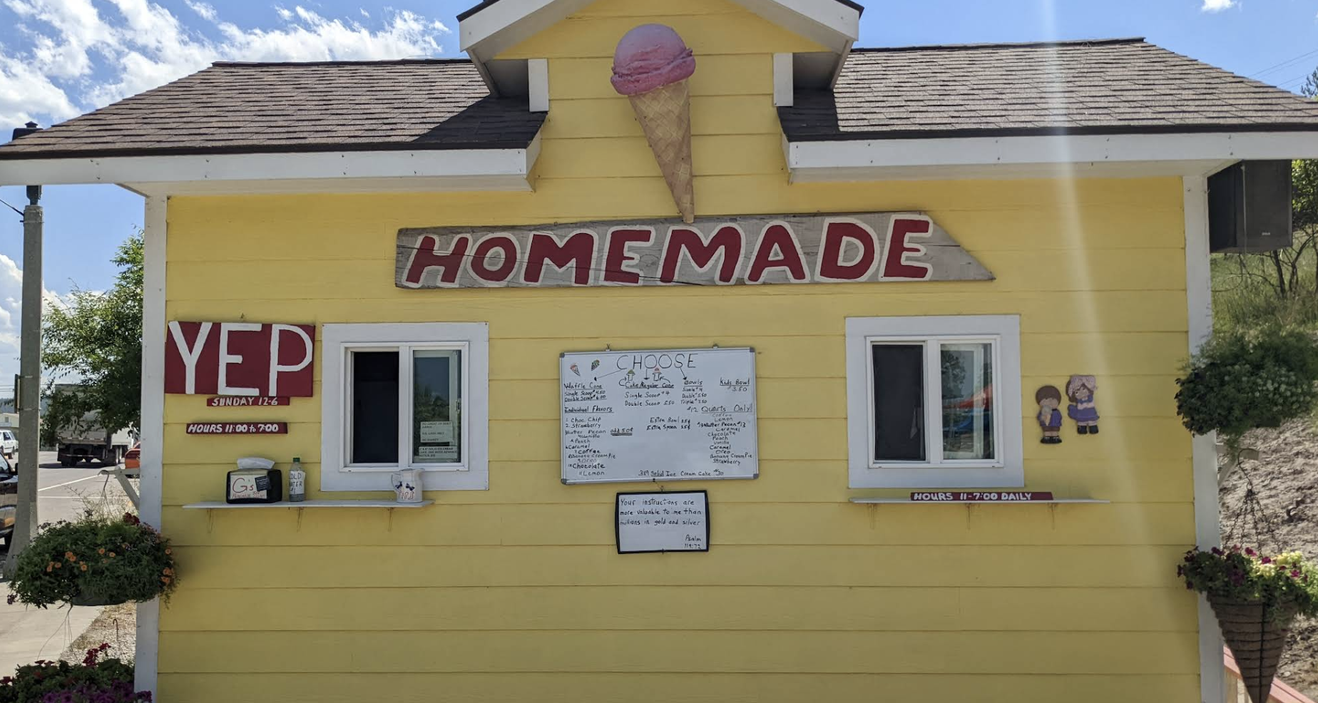 Yellow homemade ice cream stand with a pink ice cream cone sign on top, red and white signage, and small cartoon figures on the right side.