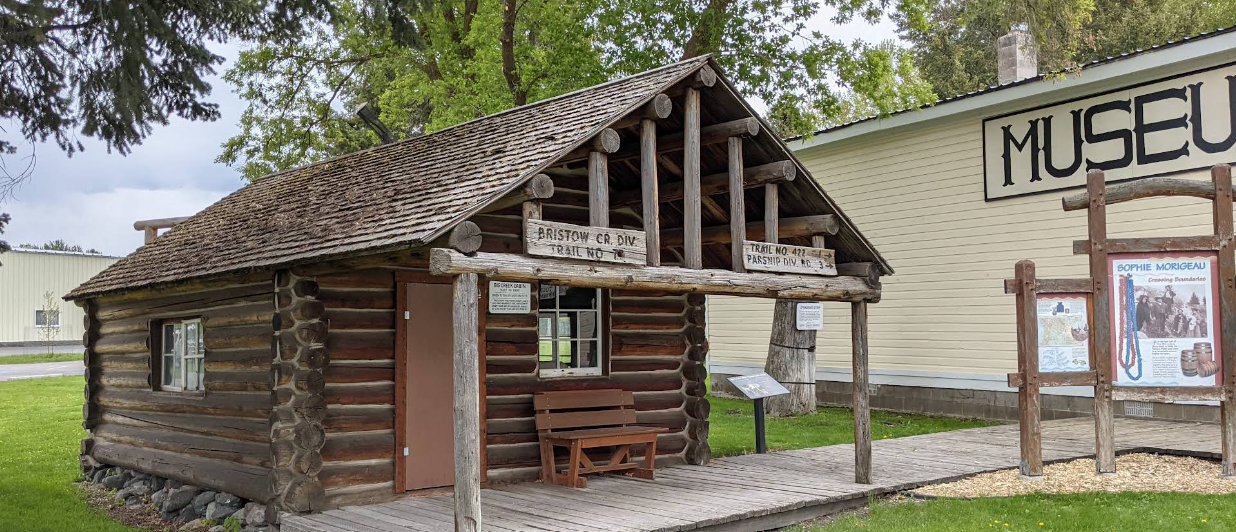 A small log cabin museum with a wooden porch, located outside with trees and grass nearby, and a larger building labeled 'Museum' in the background.