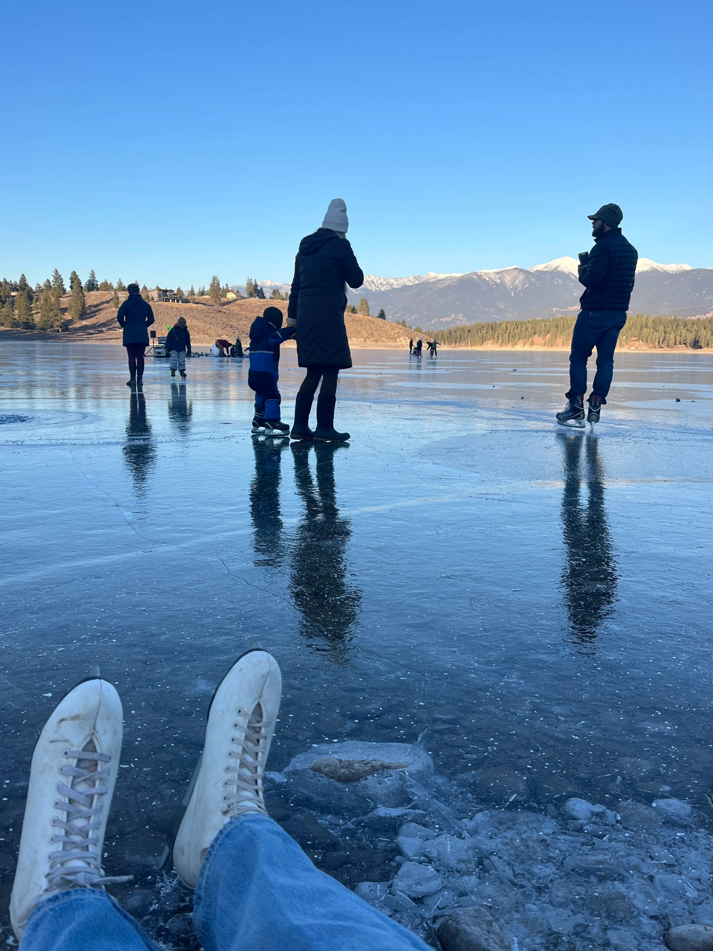 Person relaxing on icy lake with snow-capped mountains in the background, other people ice skating and taking photos, wearing winter clothing.
