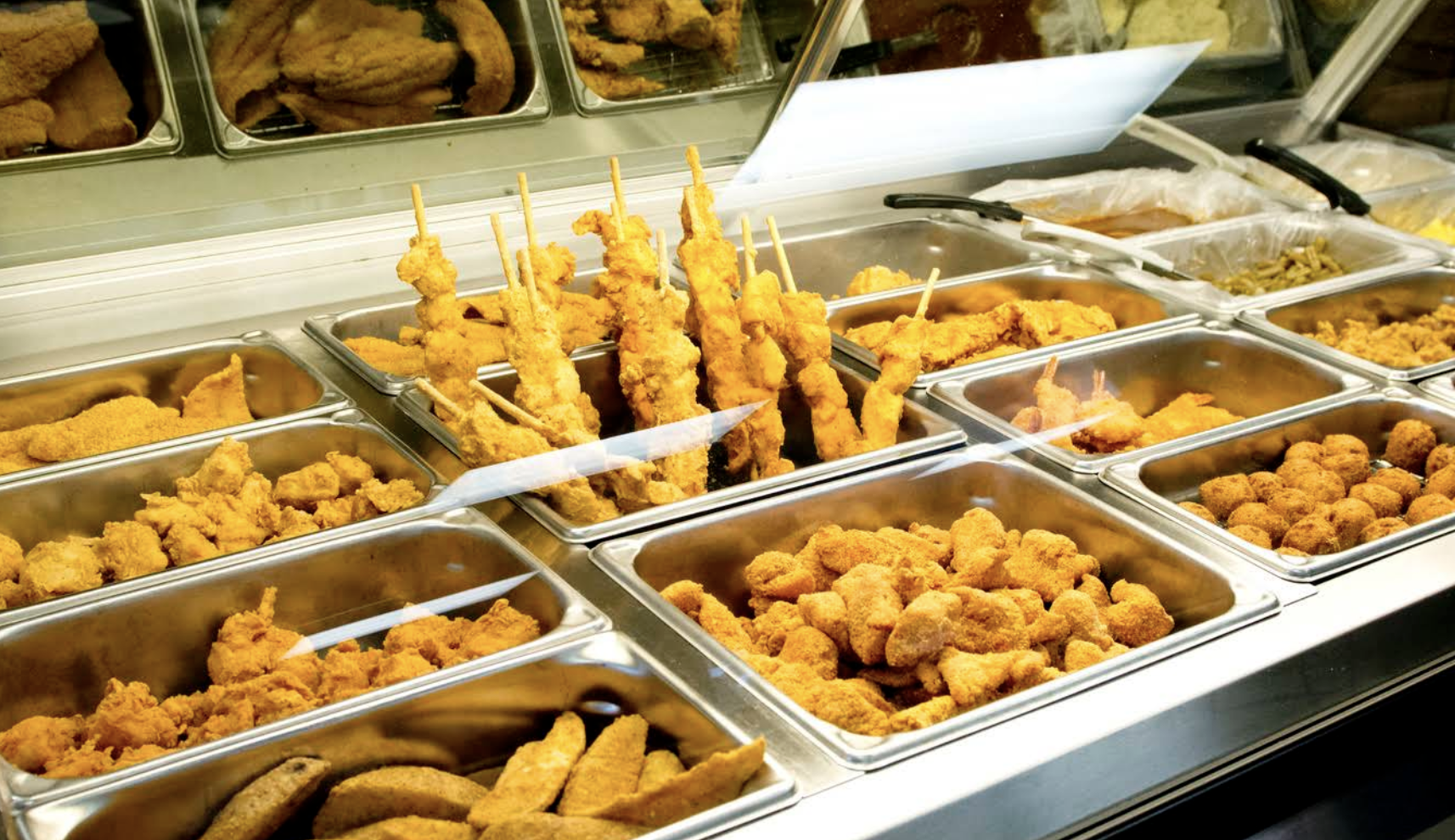 Display case of various fried foods including fried chicken tenders, fried chicken wings, chicken skewers, chicken nuggets, and other fried items in metal trays at a restaurant or buffet.