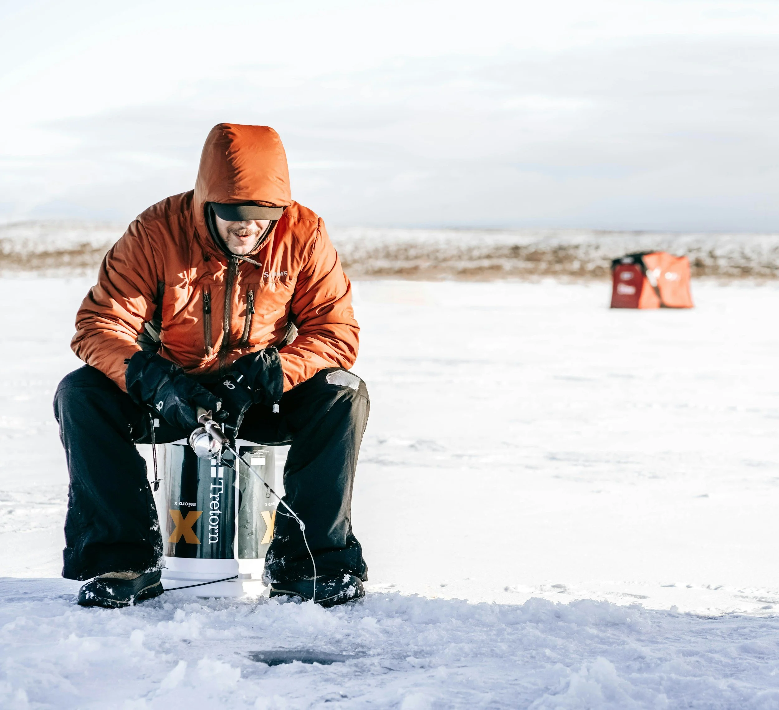 A person dressed in an orange jacket, black pants, and a black cap, sitting on a snow-covered surface, with a large cylindrical container marked "X" in front of them, and a red bag in the background, outdoors in a winter landscape.