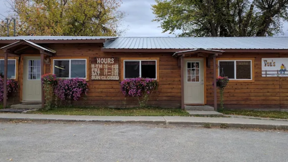 A wooden building with a metal roof, displaying business hours sign and pink floral arrangements outside, with a sidewalk and street in front.