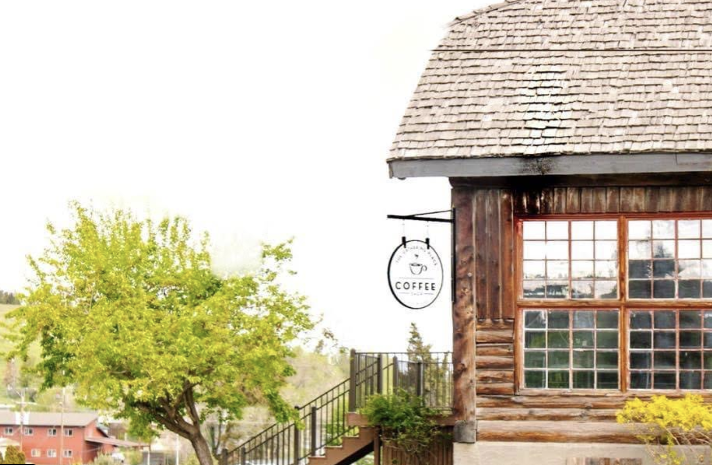 A rustic coffee shop with a hanging sign and large windows, surrounded by green trees and a clear sky.