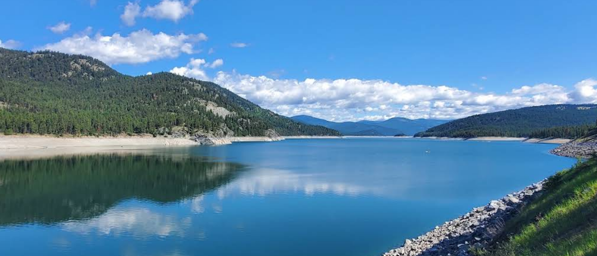 A serene mountain lake with calm blue water, surrounded by forested hills and mountains under a partly cloudy sky.