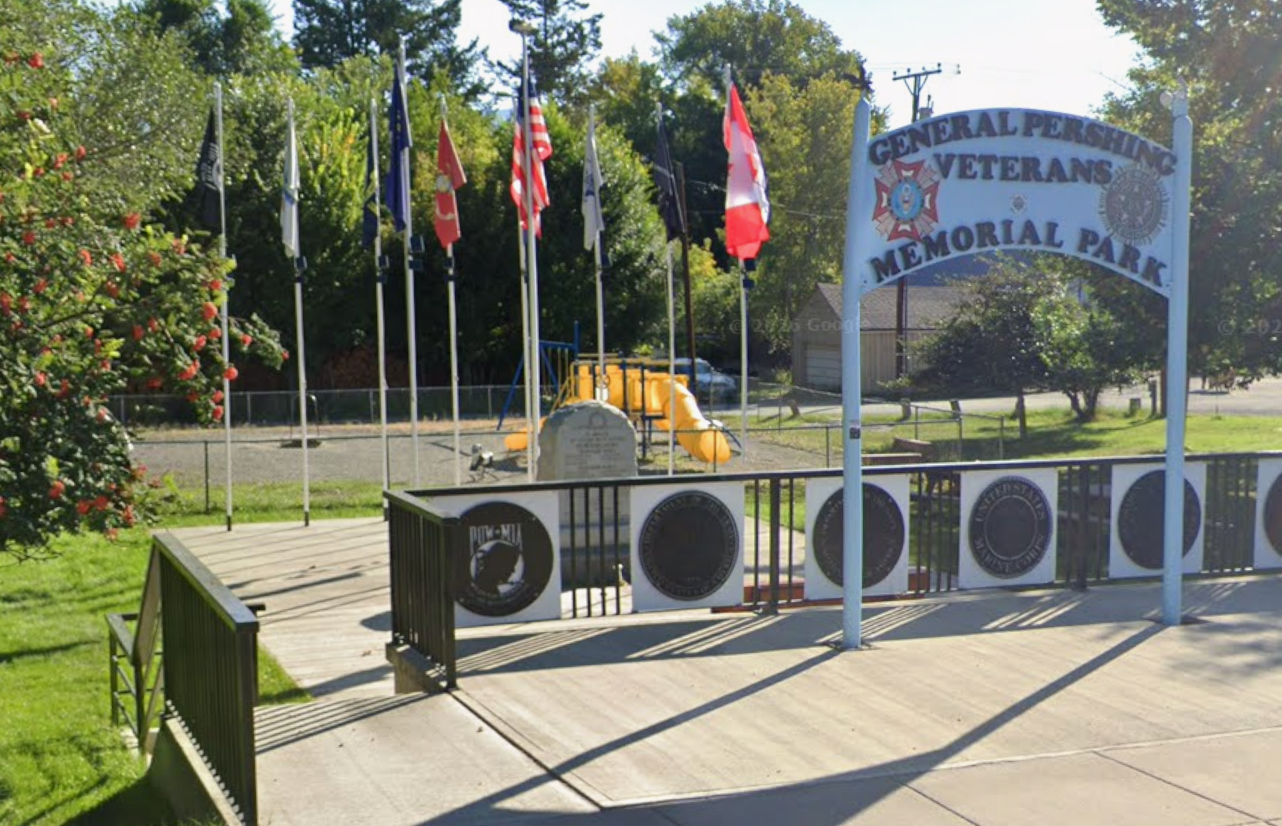 A memorial park with a sign that reads 'General Pershing Veterans Memorial Park' surrounded by flagpoles with various flags, a playground, trees, and a grassy area.