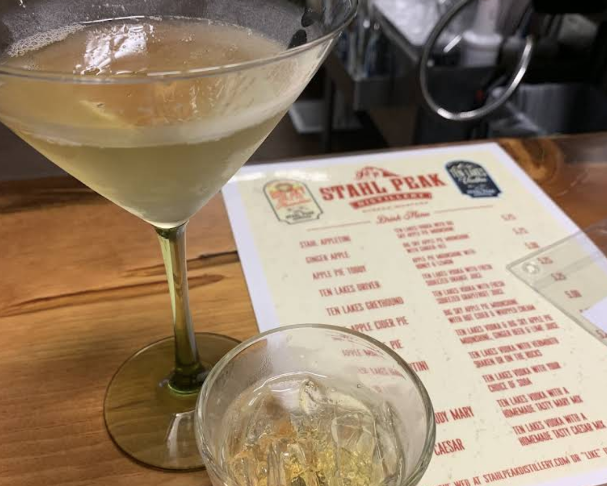 A martini glass with a light-colored cocktail on a wooden bar or table, with a menu in the background.