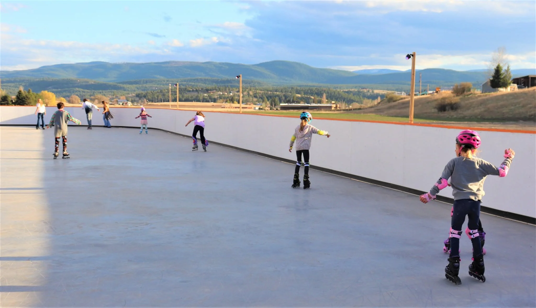Children ice skating outdoors on an ice rink with mountains and blue sky in the background.