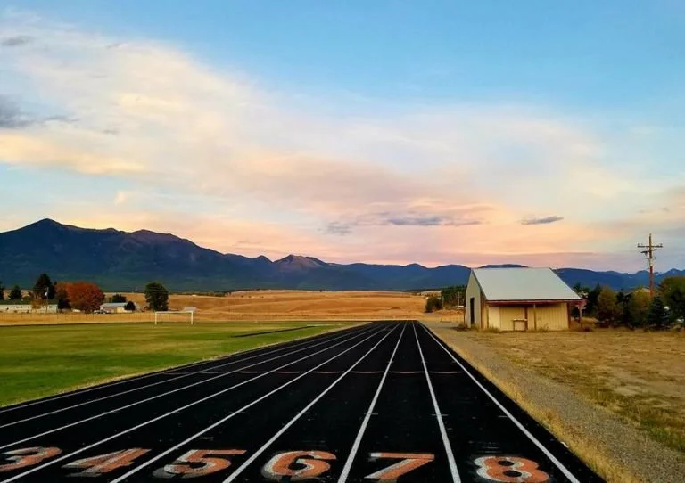 A running track with numbered lanes, a green field on the left, a barn and utility pole on the right, and mountains in the distance under a partly cloudy sky during sunset.