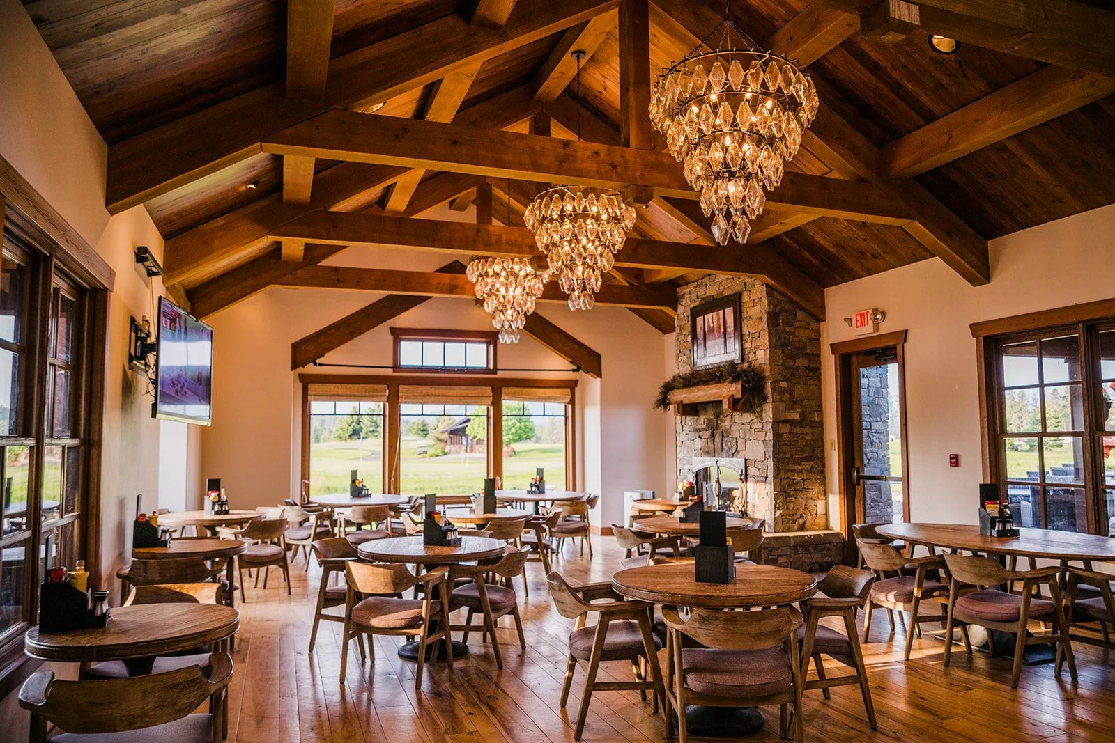 Interior of a rustic-style restaurant with wooden tables and chairs, large windows with views of green landscape, stone fireplace, wooden beams in the ceiling, and chandeliers hanging from the ceiling.