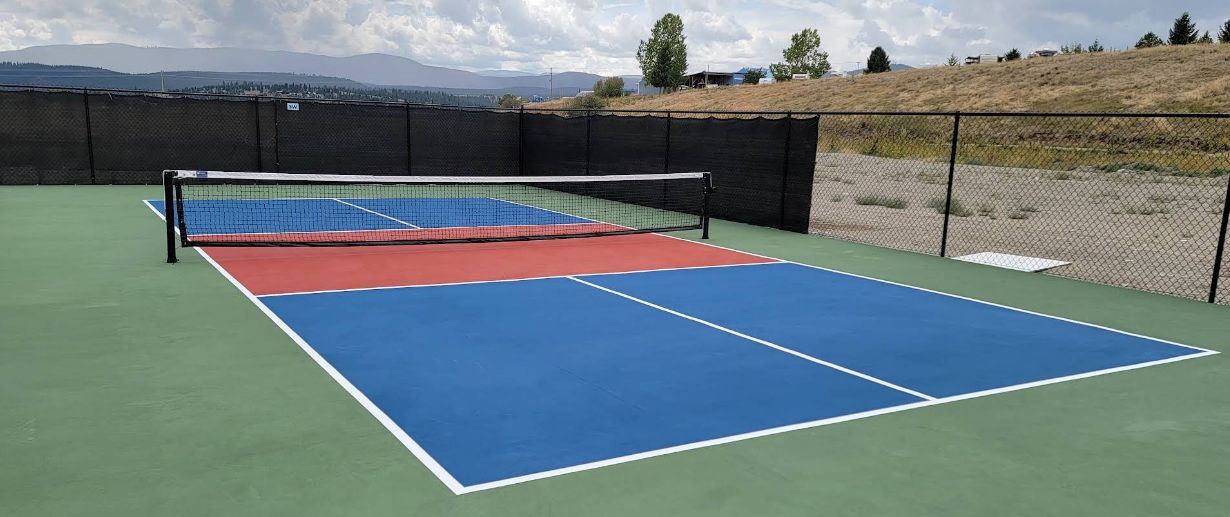 An outdoor pickleball court with a net, painted in blue, red, and green, enclosed by black windscreen fencing, with a grassy hillside and cloudy sky in the background.