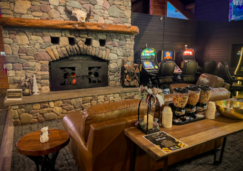 Cozy indoor scene with a stone fireplace, leather couches, and a variety of cereals and snacks on a wooden table in front of the couches, with gaming machines in the background.