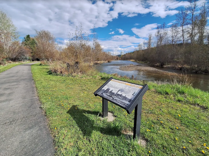 A scenic riverside park with a walking path, a grassy area, and a sign explaining the history or ecology of the area, under a partly cloudy sky during daytime.