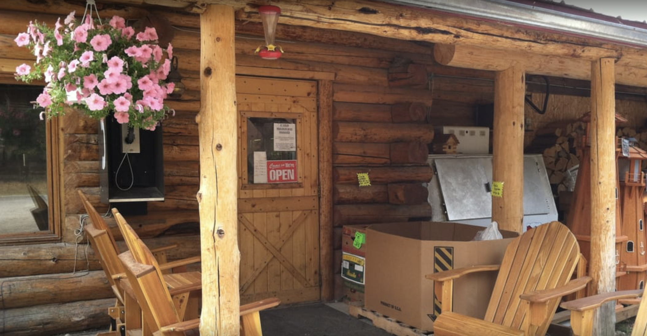 A rustic wooden log cabin porch with pink petunias hanging in a black container near the window, wooden chairs, a cardboard box, a closed door with a small window and an open 'Open' sign, logs stacked against the wall, and some equipment and signs in the background.