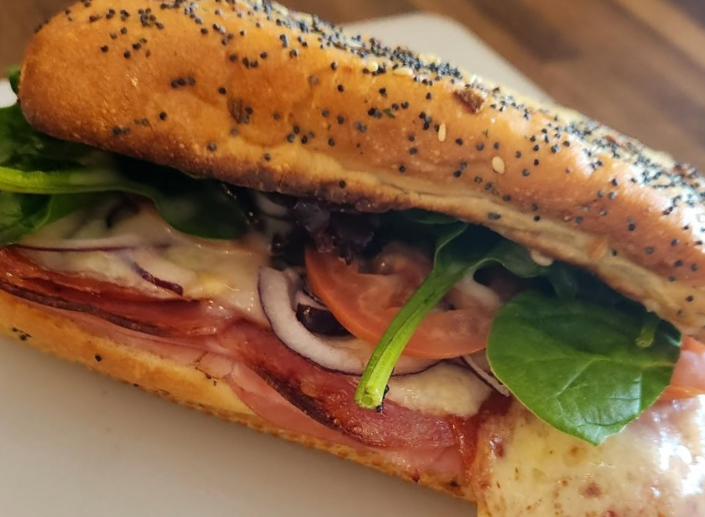 Close-up of a sandwich with poppy seed bread, filled with lettuce, tomato, onion, meat, cheese, and other sandwich ingredients.