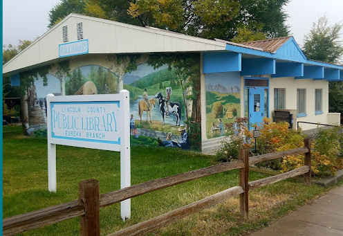 A mural of a rural scene with horses on the side of a building, outside Lincoln County Public Library, Cherokee Branch, with a sign and a wooden fence in front.