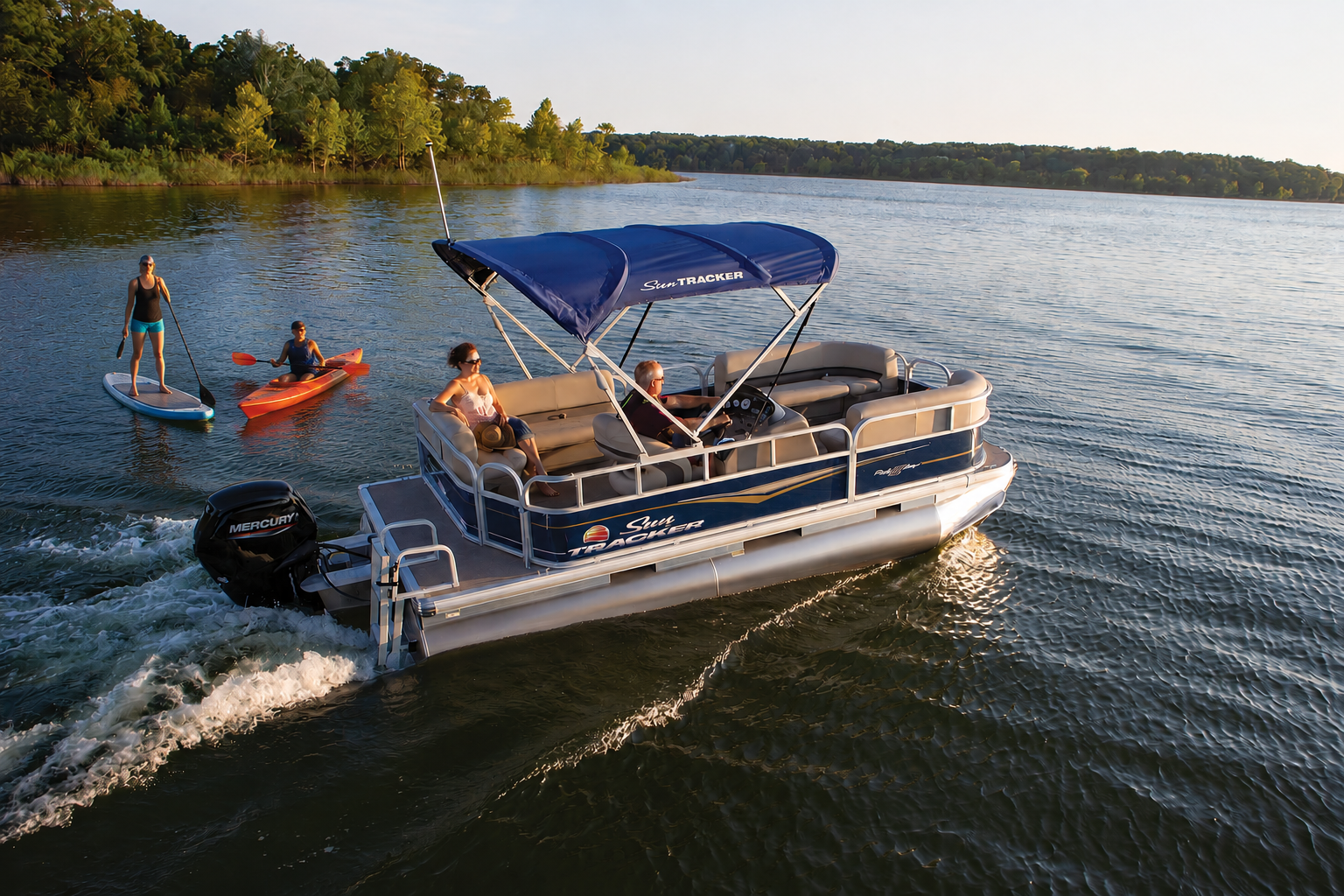Family riding in a pontoon boat on a lake with two children kayaking nearby, surrounded by trees.