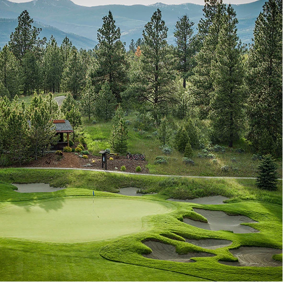 A golf course with a green, sand traps, and a flag, surrounded by trees and mountains in the background.