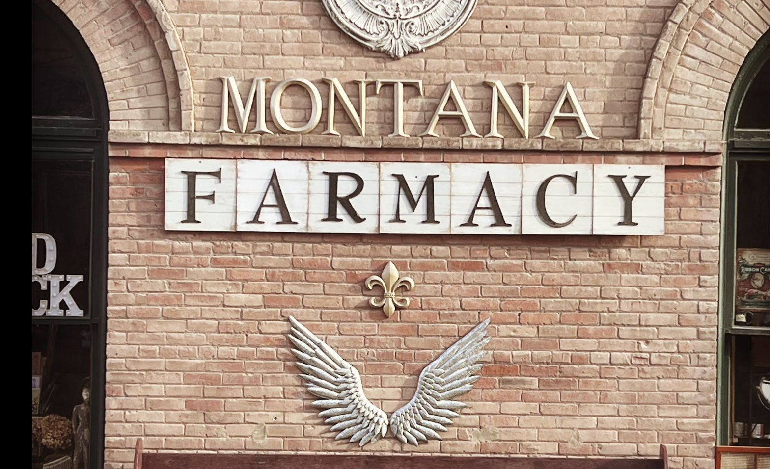 Sign for Montana Pharmacy on a brick wall, with a decorative fleur-de-lis and angel wings below.