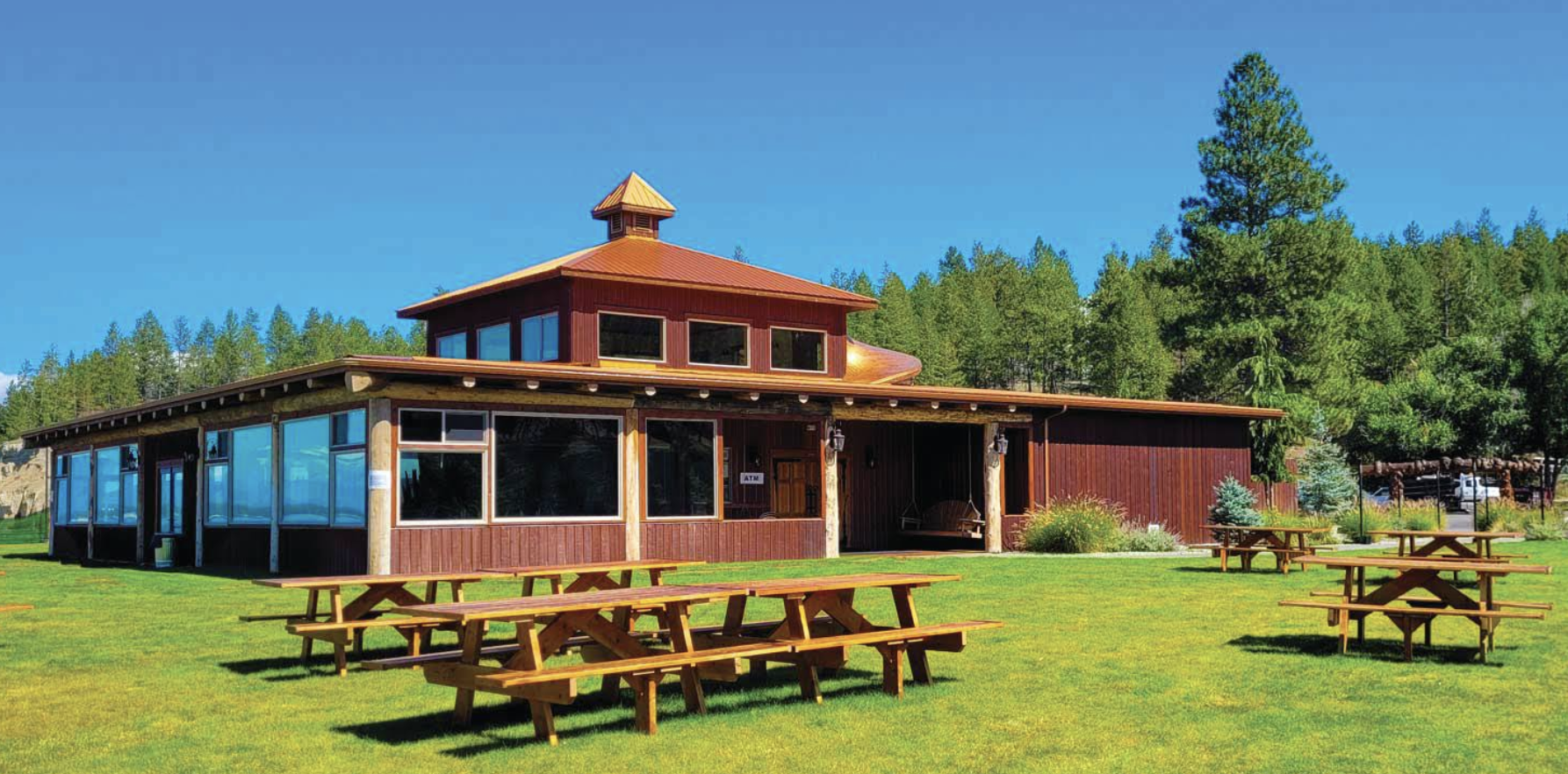 A rustic building with large windows, wooden exterior, and a small tower, situated in a lush green lawn with picnic tables, surrounded by pine trees under a bright blue sky.