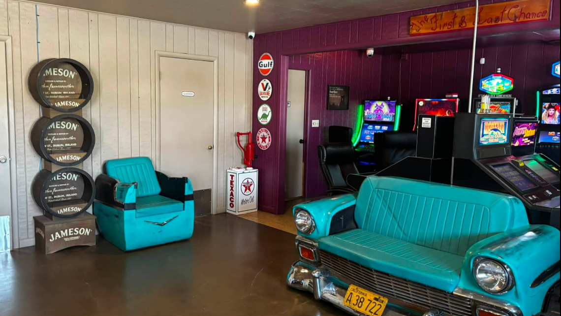 Interior view of a retro arcade with vintage car-themed seating and slot machines, orange sign on ceiling reads 'First & Last Chance'.