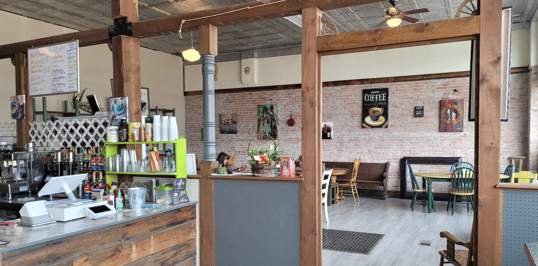 Interior of a cozy coffee shop with rustic brick walls, wooden beams, and a variety of seating options including tables, chairs, and a bench. Decor includes coffee-themed artwork, a menu board, and a small counter with coffee-making equipment and cups.