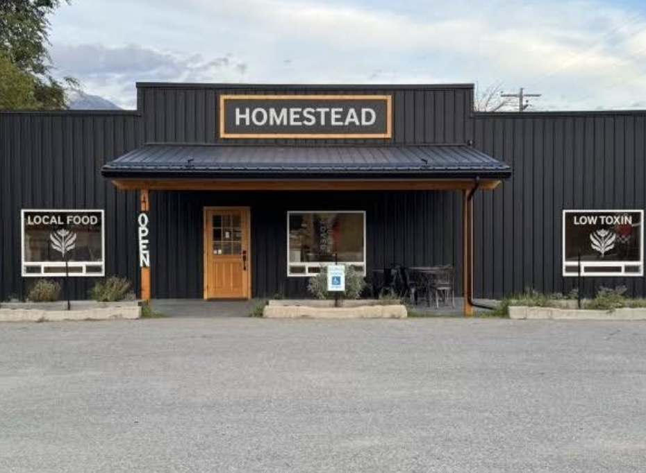 Black storefront with orange sign reading 'HOMESTEAD', labeled as a place with local food and low toxin options. Features a wooden door, large window, outdoor seating, and a sign indicating 'OPEN'.