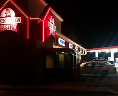Nighttime view of a casino with illuminated signs, including a poker sign, and parked cars outside.
