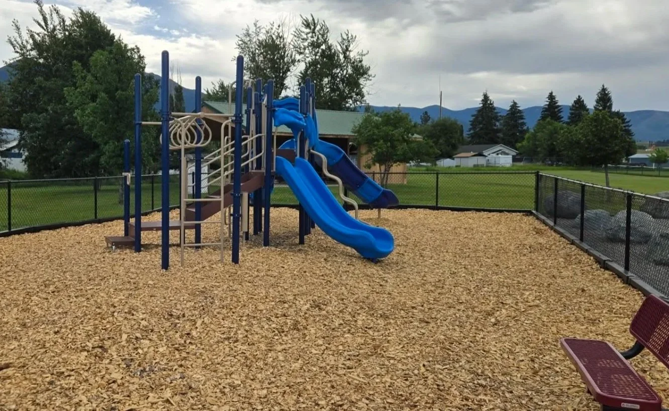 Empty playground with a blue slide and climbing structure, wood chip ground cover, fenced yard, trees, and mountains in the background under cloudy sky.