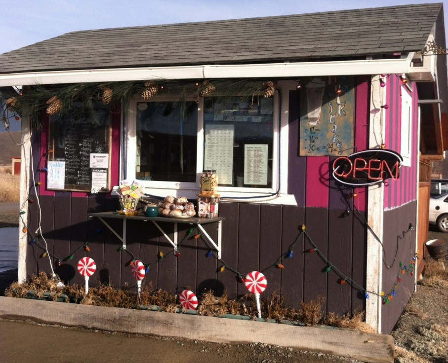 A small shop or food stand decorated with Christmas lights and ornaments, featuring a pink and brown exterior with a window, an "OPEN" sign, and holiday decorations including peppermint candy decorations and pine garland.