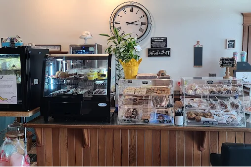 Pastry display case with cookies and baked goods at a bakery counter, with a large wall clock, potted plant, and food menu signs in the background.