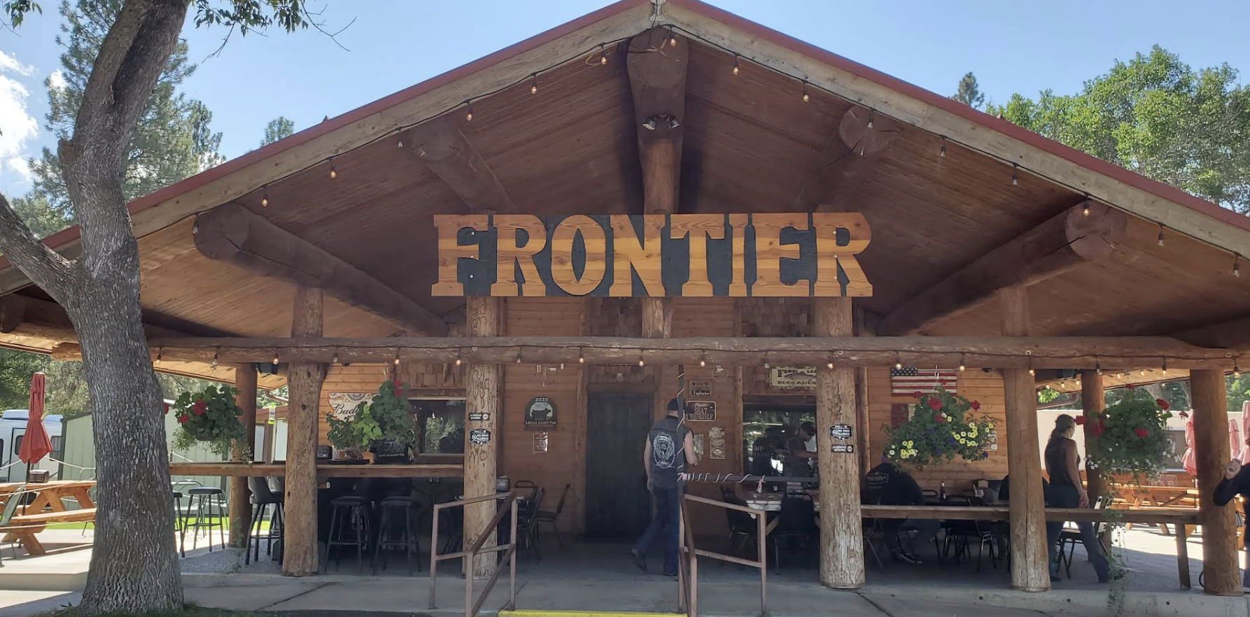 Frontier building with wooden logs, hanging string lights, and a large sign that says 'FRONTIER'. There are hanging flower baskets, people inside and outside, and trees surrounding the area.